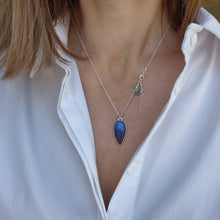 Load image into Gallery viewer, A person wearing a white shirt is showcasing a silver pendant with a blue labradorite stone and a small silver charm attached to it. The pendant comes with an adjustable chain.